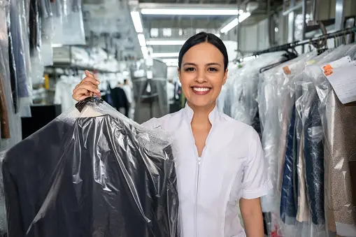 Beautiful employee at a laundromat holding a dry cleaned outfit covered with a bag while facing camera smiling very happy
